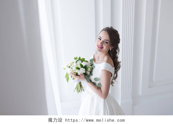 在白色背景墙前的开心的新娘快乐的年轻新娘摆在优雅的白色礼服与婚礼花束